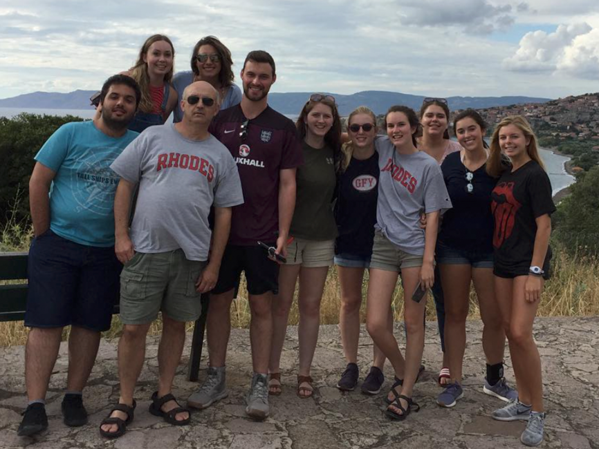 Photo of students and one male professor in shorts and t-shirts in front of water and hills in Greece