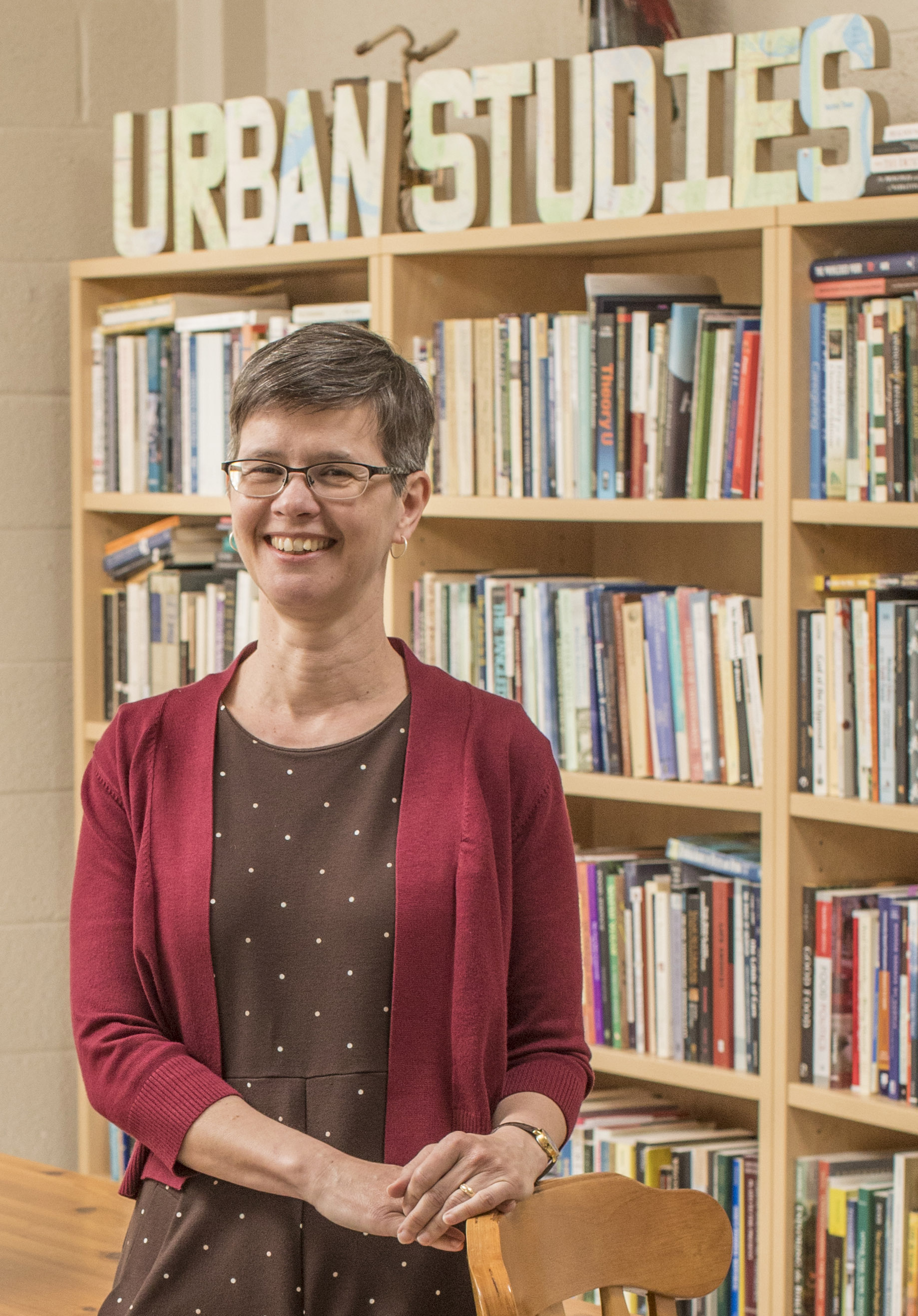 a woman with short hair stands in front of a bookcase with the words Urban Studies on top