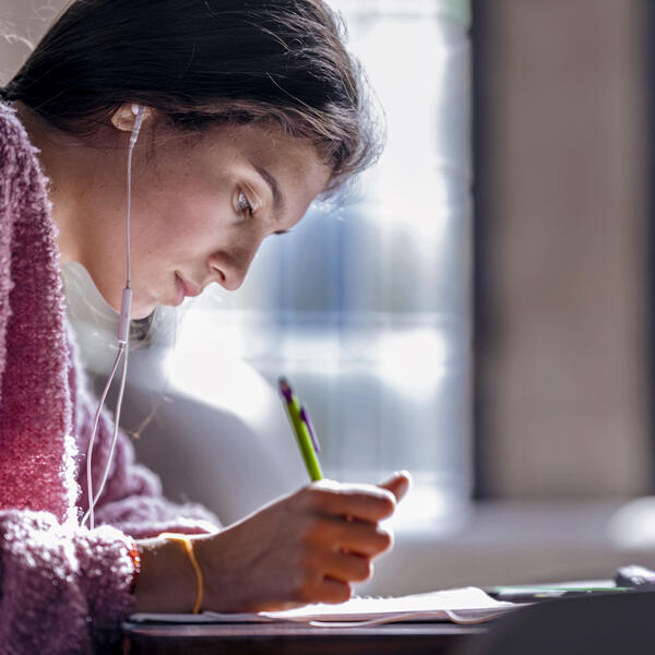 A young woman with dark hair and a pink sweater looks down while writing as light streams through the windows behind her.