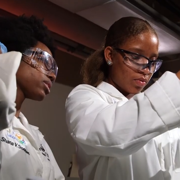 Two women in lab coats, protective eyewear, and gloves transfer liquid into a beaker.