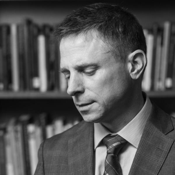 A professor in suit and tie stands in front of his bookcase.