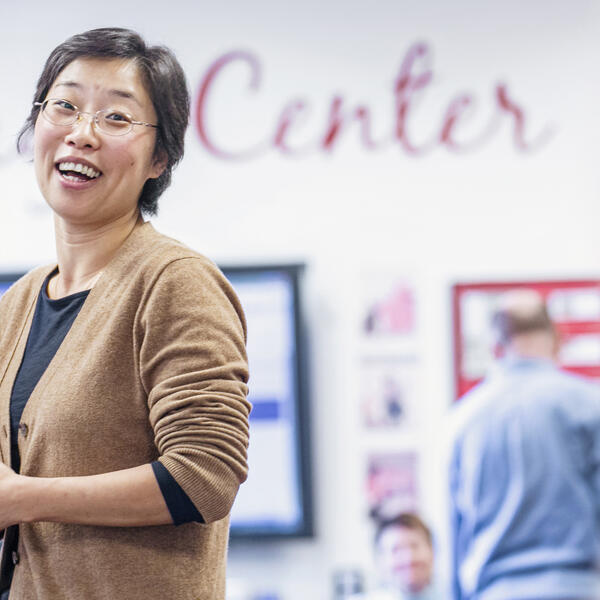 An Asian woman in a tan sweater laughs with students