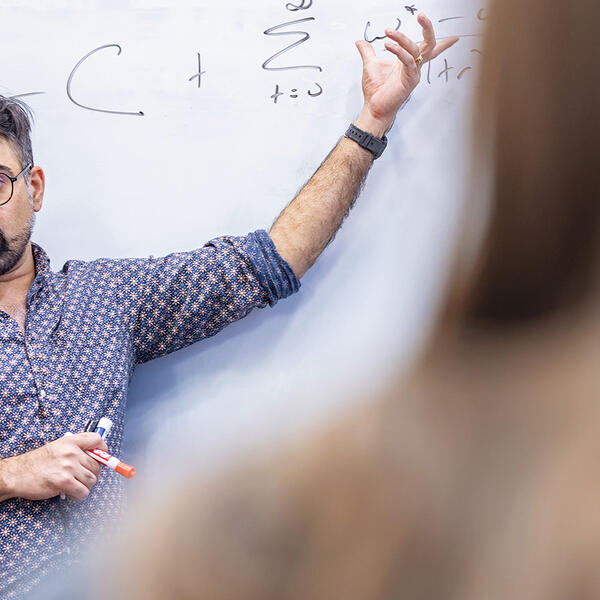 a male professor gestures at a whiteboard with equations