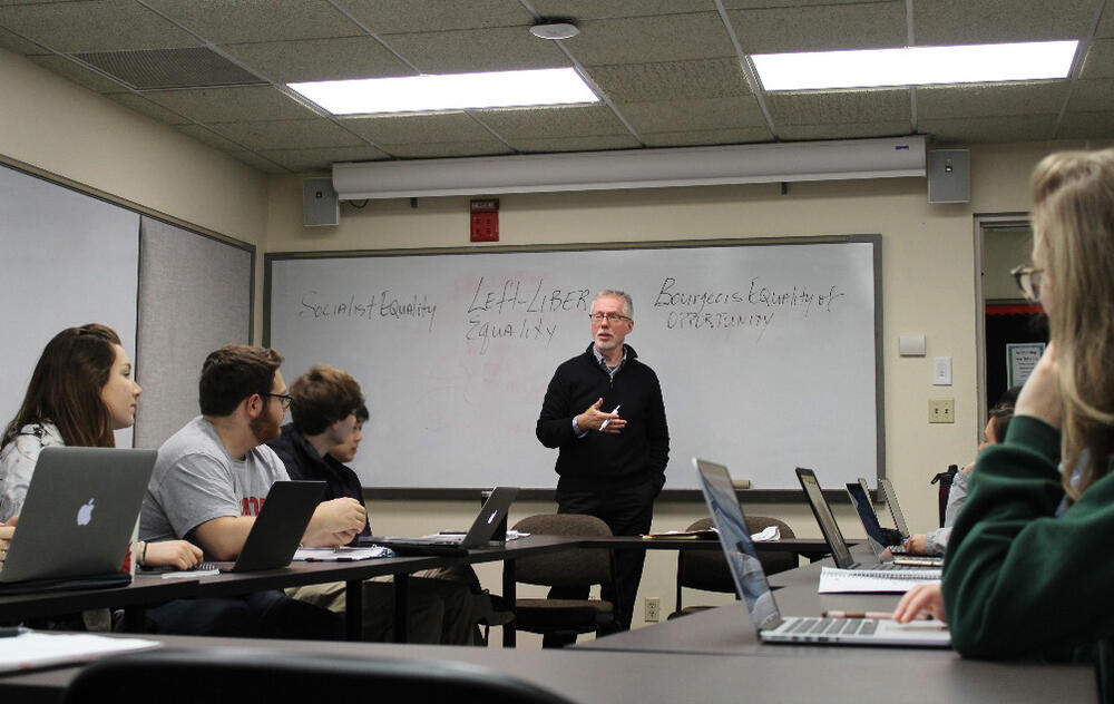 a male professor talks in front of a whiteboard
