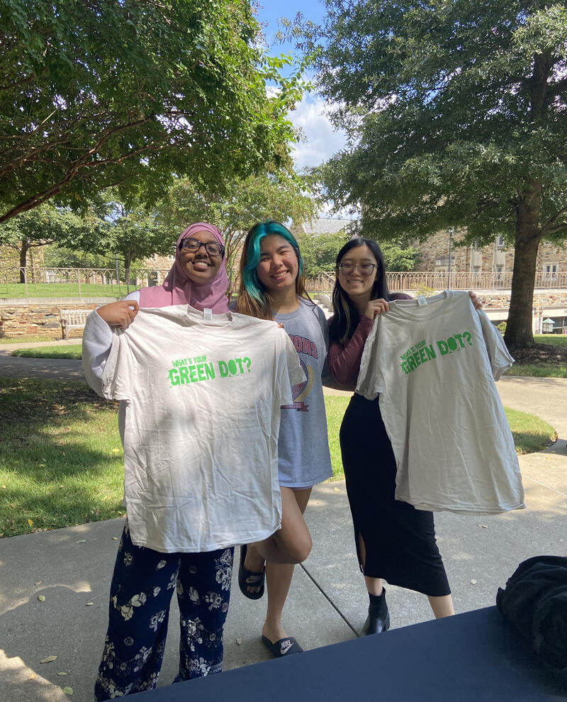 three students hold up t-shirts that say "What's your green dot?"